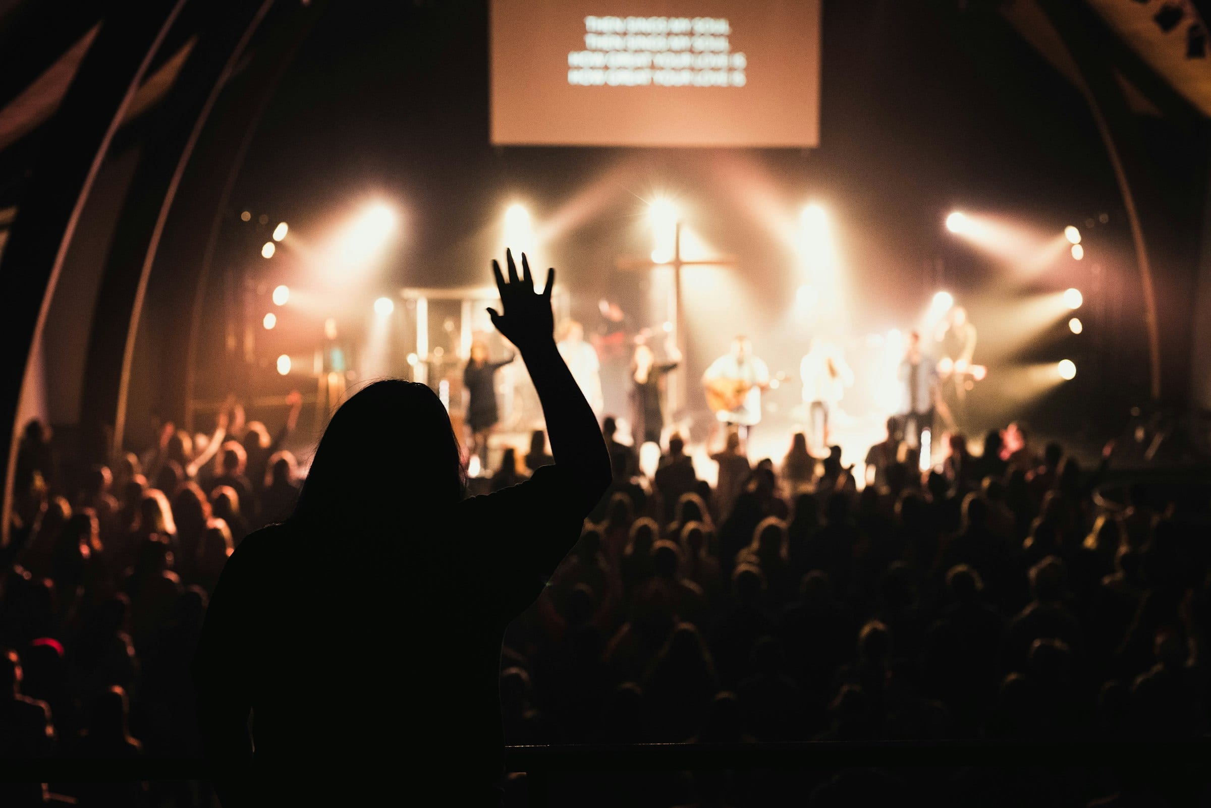 A worship service seen from the back of the room, one hand raised toward the stage lights
